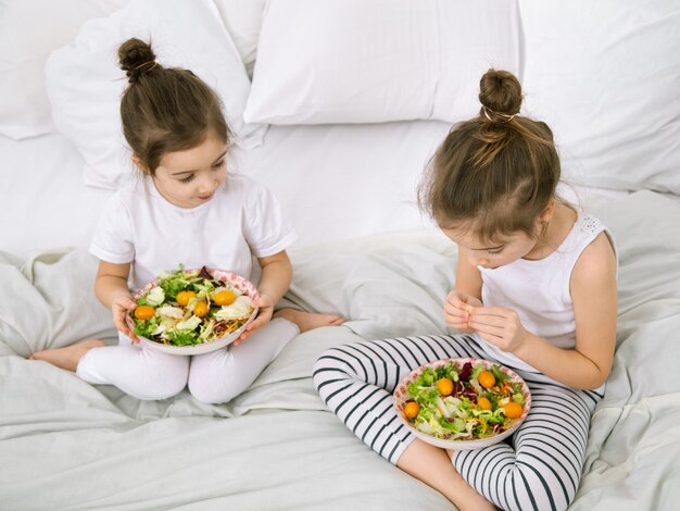 Comida saudável em casa. Felizes dois filhos bonitos comendo frutas e legumes no quarto na cama. Alimentação saudável para crianças e adolescentes.