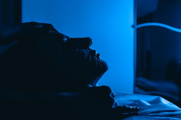 Composição escura abstrata com iluminação azul. a jovem está deitada em uma máscara hidratante de tecido.