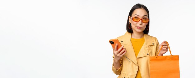 Compradora de menina asiática elegante usa óculos escuros segurando sacola de compras e smartphone indo para descontos em lojas em cima de fundo branco