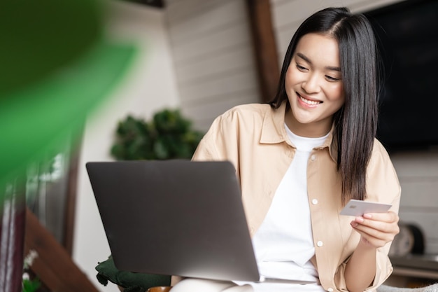 Compras na internet, jovem mulher asiática, pagando uma compra online usando cartão de crédito e laptop sentado ...
