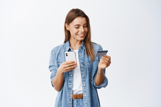 Compras on-line mulher bonita sorridente pagando pelo pedido usando cartão de crédito plástico para pagar com o celular em pé contra o fundo branco