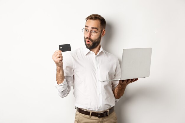 Compras online. Empresário surpreso segurando laptop, parecendo impressionado com o cartão de crédito, em pé