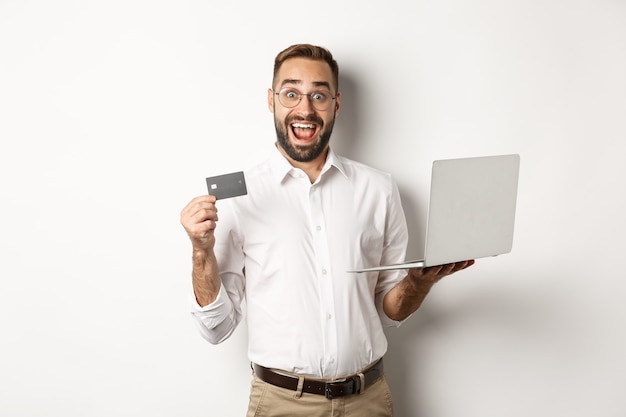 Compras online. Homem bonito, mostrando o cartão de crédito e usando o laptop para fazer um pedido na internet, em pé