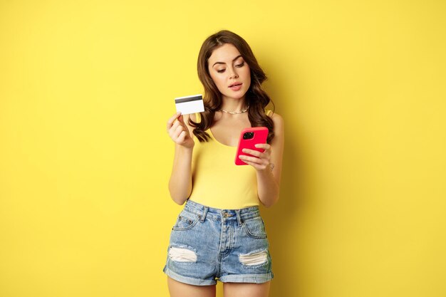 Compras online. Mulher morena elegante segurando smartphone e cartão de crédito, pagando no aplicativo, usando o aplicativo de celular, comprando smth, de pé sobre fundo amarelo