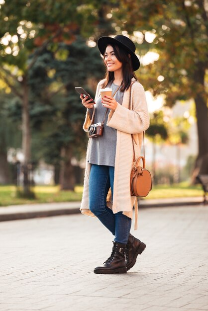 Comprimento total da mulher asiática feliz no desgaste elegante, segurando o telefone móvel e a xícara de café em pé no parque ao ar livre