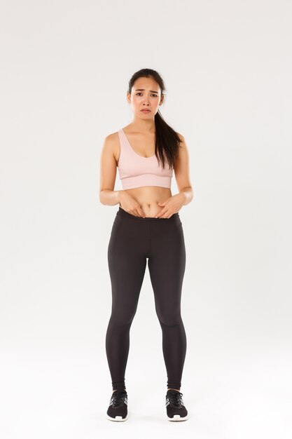 Comprimento total de menina asiática chateada e sombria em roupas de fitness, mostrando gordura na barriga, reclamando do corpo, carrancuda e parecendo desapontada, começando o treino, tentando perder peso, fundo branco.