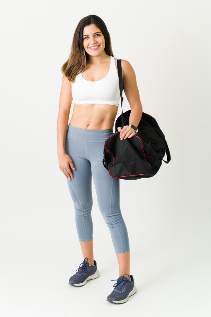 Foto grátis comprimento total de uma atleta feminina atraente vestindo roupas esportivas sorrindo enquanto carregava uma bolsa de ginástica. mulher jovem confiante com abs tonificado