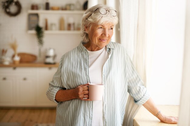 Conceito de aconchego, domesticidade e lazer. Retrato de uma elegante mulher de cabelos grisalhos com óculos redondos na cabeça, apreciando o café da manhã, segurando uma caneca, olhando para fora através do vidro da janela