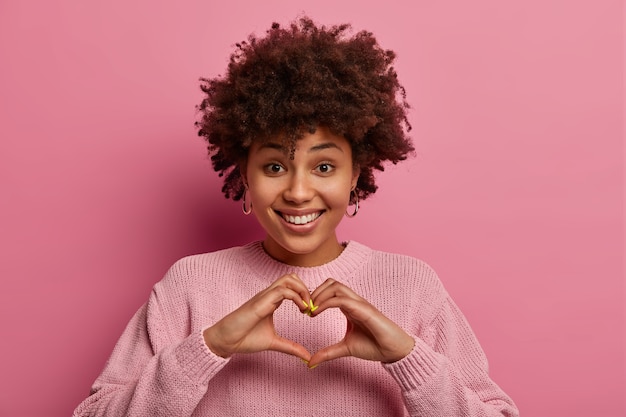 Foto grátis conceito de afeto e relacionamento. ainda bem que a mulher étnica encantada molda o coração com as mãos, sorri positivamente, demonstra o símbolo do amor, usa um suéter rosa pastel, faz gestos internos. foto monocromática