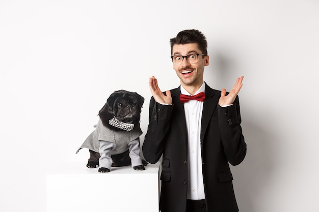 Conceito de animais, festa e celebração. Homem bonito e cachorro fofo em trajes de terno olhando surpreso para a câmera, reagindo na oferta promocional com um fundo branco espantado