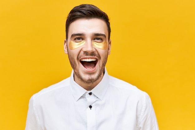 Conceito de beleza e cuidados com a pele. Foto de um jovem rapaz animado e alegre com a barba por fazer com manchas douradas sob os olhos para parecer acordado e revigorado, gritando de excitação, abrindo a boca bem aberta