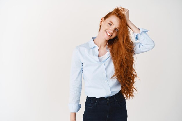 Foto grátis conceito de beleza e cuidados com os cabelos. mulher jovem com olhos azuis e sardas tocando um cabelo vermelho brilhante e saudável e sorrindo na frente, na parede branca