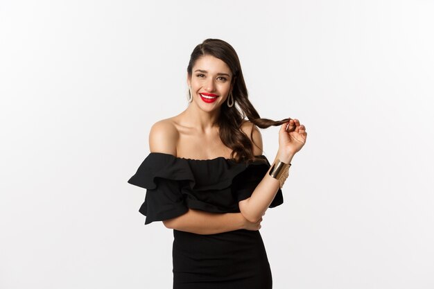 Conceito de beleza e moda. Glamour mulher de vestido preto, lábios vermelhos, brincando com o cabelo e ouvindo, olhando para a câmera com interesse, em pé sobre um fundo branco.