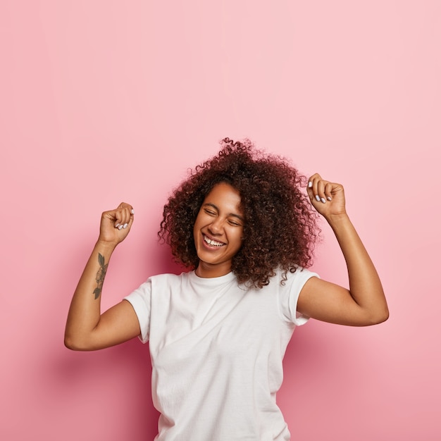 Conceito de bem-estar e alegria. foto vertical de mulher encaracolada despreocupada e satisfeita levanta os braços tatuados, sente-se relaxada e feliz, move-se ao ritmo de uma música agradável, tem os olhos fechados, dança ativamente