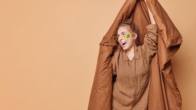 Conceito de bom dia uma jovem alegre e adorável vestida de pijama segura cobertor aplica manchas verdes de hidrogel sob os olhos para danças de tratamento de pele contra fundo marrom simulado espaço