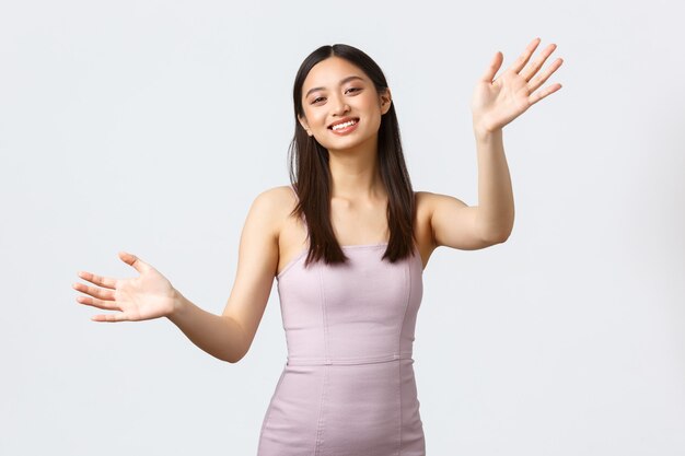 Conceito de celebração, beleza e glamour. Encantadora e bela anfitriã feminina asiática da festa em um vestido de noite luxuoso, cumprimentando os hóspedes, estendendo as mãos para um abraço e sorrindo com fundo branco.