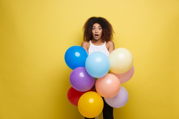 Conceito de Celebração - Close up Retrato feliz jovem mulher africana bonita em t-shirt preto sorrindo com balão de festa colorida. Fundo amarelo do estúdio Pastel.