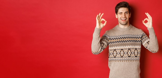 Conceito de celebração do Natal, férias de inverno e estilo de vida. Homem bonito confiante e atrevido, garantia tudo de bom, dando sinais de ok e piscando para a câmera, fundo vermelho