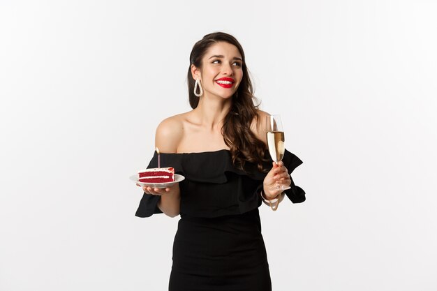 Conceito de celebração e festa. Mulher elegante segurando bolo de aniversário com vela e bebendo champanhe, sorrindo e olhando para o lado, em pé sobre um fundo branco.