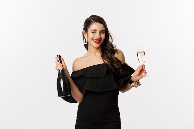 Conceito de celebração e festa. Mulher morena elegante com vestido glamour, segurando a garrafa e a taça de champanhe, se divertindo no feriado de ano novo.