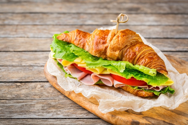 Conceito de comida, pequeno-almoço, manhã e almoço. sanduíche de croissant fresco com presunto, queijo, alface e tomate em uma mesa de madeira