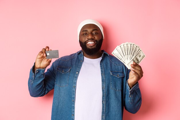 Conceito de compras. Bonito jovem negro mostrando o cartão de crédito do banco e dinheiro, sorrindo satisfeito, em pé sobre um fundo rosa.