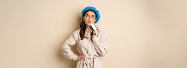 Conceito de compras e descontos Jovem garota moderna elegante posando com cartão de crédito parecendo feliz e sorrindo em pé sobre fundo bege