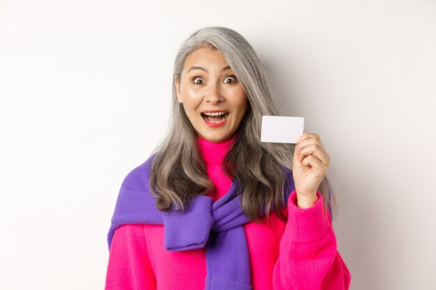 Conceito de compras. Feliz asiático velho aldy parecendo impressionado e mostrando o cartão de crédito de plástico do banco dela, em pé sobre um fundo branco.