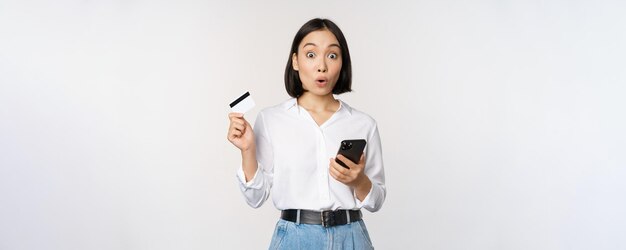 Conceito de compras on-line Imagem de menina asiática surpresa segurando cartão de crédito e smartphone olhando espantado em descrença no fundo branco da câmera