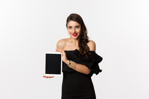 Conceito de compras online. Mulher bonita tentada em um vestido preto, mostrando a tela do tablet digital, em pé sobre um fundo branco. Copie o espaço