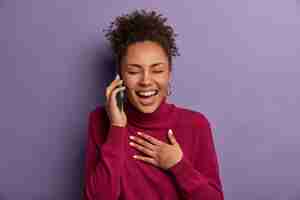 Foto grátis conceito de comunicação, pessoas e tecnologia. mulher afro-americana ri despreocupada enquanto fala no celular, fecha os olhos e não para de rir, ouve algo engraçado ou muito positivo de um amigo