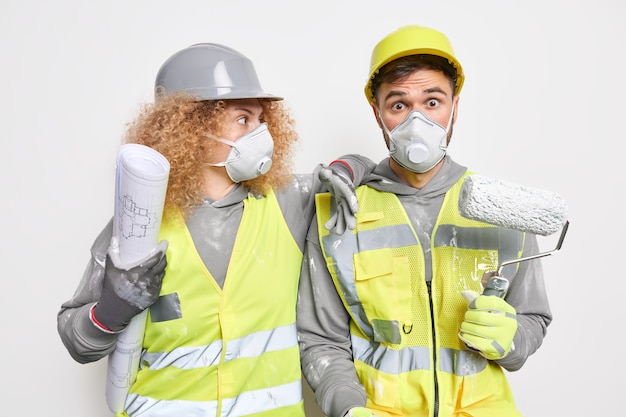 Conceito de construção e engenharia de reparação. Equipe de trabalhadores de manutenção chocados, vestidos com roupas de trabalho, segurando a planta do papel e pintando com rolo de pintura na parede interior do projeto