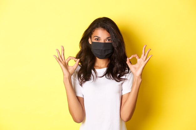 Conceito de coronavírus, pandemia e estilo de vida. Retrato de uma garota afro-americana confiante, usando máscara preta para se proteger de covid-19, mostrando sinais de ok, fundo amarelo.