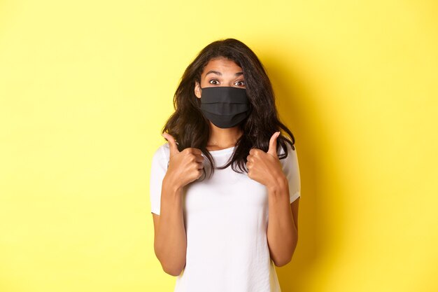 Conceito de coronavírus, pandemia e estilo de vida. Retrato de uma linda garota afro-americana na máscara facial preta, mostrando o polegar para cima em aprovação, como algo bom, fundo amarelo.