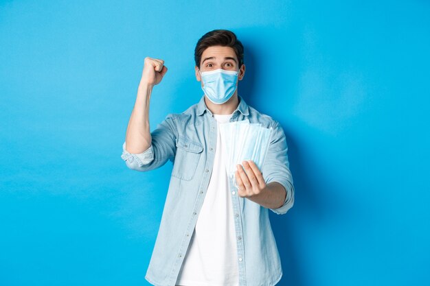 Conceito de coronavírus, quarentena e distanciamento social. Homem alegre mostrando máscaras médicas e fazendo o punho bombar, celebrando ou triunfando, em pé contra um fundo azul