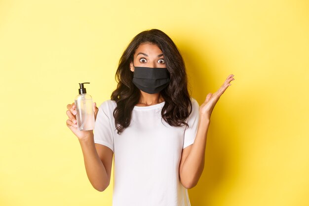 Conceito de covid-19, distanciamento social e estilo de vida. Imagem de mulher afro-americana animada, usando máscara facial, levantando as mãos surpreso, segurando um desinfetante para as mãos, fundo amarelo.