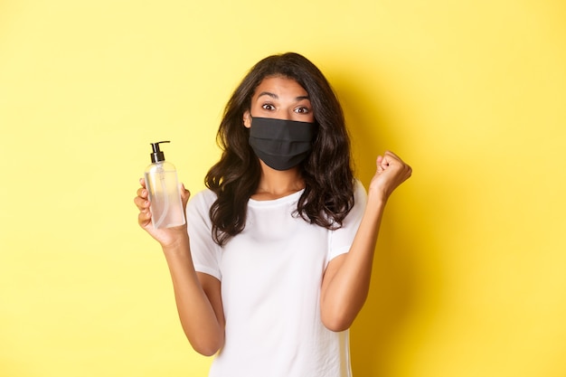 Conceito de covid-19, distanciamento social e estilo de vida. Imagem de uma mulher afro-americana feliz com uma máscara facial, sentindo-se feliz por ter encontrado um bom desinfetante para as mãos, regozijando-se com o fundo amarelo