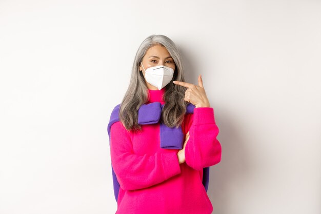 Conceito de covid, pandemia e distanciamento social. Mulher asiática sênior elegante usando respirador, apontando para a máscara facial e sorrindo, em pé sobre um fundo branco.