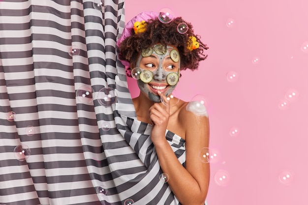 Foto grátis conceito de cuidado de pele de bem-estar de higiene de pessoas. mulher afro-americana feliz olha positivamente de lado aplica máscara de argila com fatias de pepino toma banho em barracas de banheiro atrás da cortina gosta de ducha