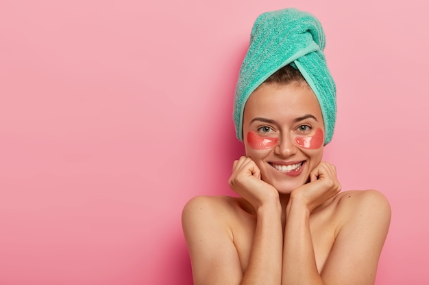Conceito de cuidados com a pele e cosmetologia. Mulher adorável e feliz aplica adesivos sob os olhos após o banho, morde os lábios, mantém as mãos sob o queixo, coloca os ombros nus contra um fundo rosa