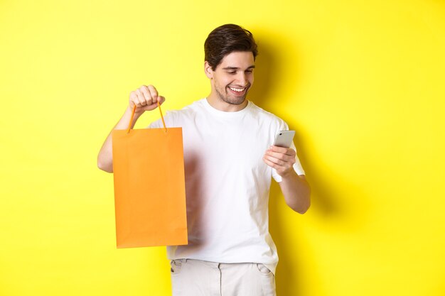 Conceito de descontos, banco online e cashback. Cara feliz, mostrando a sacola de compras e olhando satisfeito para a tela do celular, fundo amarelo.