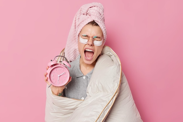 Conceito de despertar precoce. a jovem europeia com sono boceja e mantém a boca aberta segura despertador envolto em cobertor usa toalha de banho na cabeça depois de tomar banho isolado sobre fundo rosa