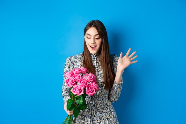 Conceito de dia dos namorados. Imagem de uma mulher jovem e atraente boquiaberta de espanto, recebendo flores surpresa, em pé sobre um fundo azul