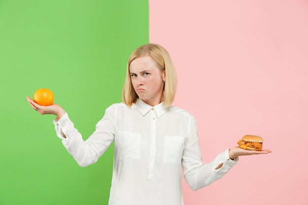 Conceito de dieta. Alimentos úteis e saudáveis. Mulher jovem e bonita escolhendo entre frutas e fast-food insalubre no estúdio. Emoções humanas e conceitos de comparação