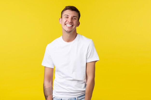 Conceito de emoções de estilo de vida, verão e pessoas. Cara caucasiano carismático bonito em camiseta branca casual, sorrindo amplamente com um sorriso branco perfeito, fundo amarelo alegre em pé.