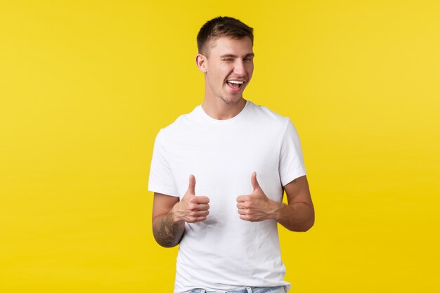 Conceito de emoções de estilo de vida, verão e pessoas. Cliente do sexo masculino insolente e satisfeito em uma camiseta branca básica, piscando e sorrindo para incentivar a experimentar, recomendar o produto com polegar para cima, fundo amarelo.