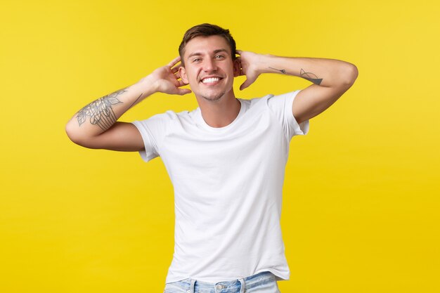 Conceito de emoções de estilo de vida, verão e pessoas. Homem feliz bonito despreocupado com sorriso branco alegre, segurando as mãos atrás da cabeça, curtindo as férias, fundo amarelo de pé.