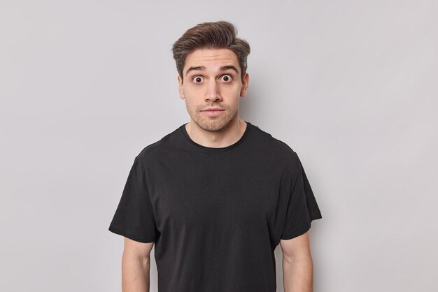 Conceito de emoções e reações de pessoas. Homem bonito atordoado olha com os olhos cheios de medo vestido de camiseta preta casual posa contra fundo branco atordoado por bancas de notícias chocantes sem palavras