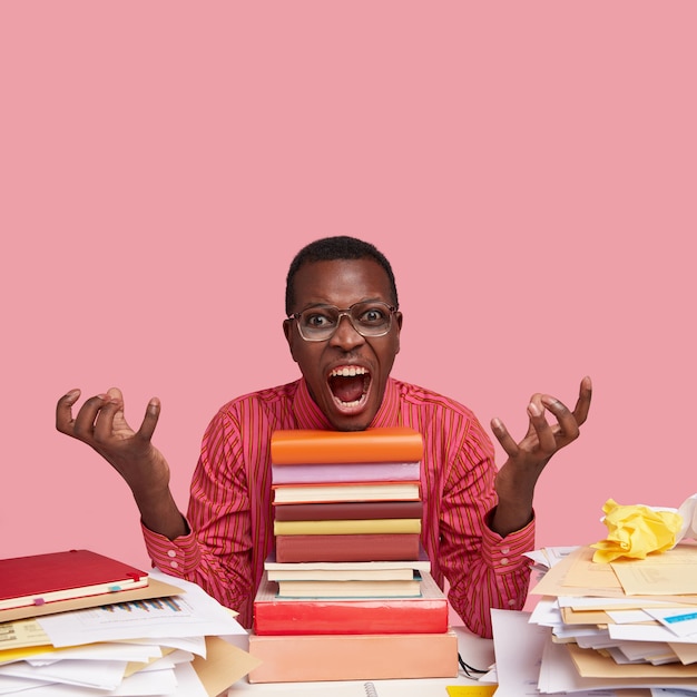 Foto grátis conceito de emoções negativas. homem jovem furioso e insatisfeito com pele escura, gesticula de raiva, abre a boca amplamente, senta-se na frente de livros