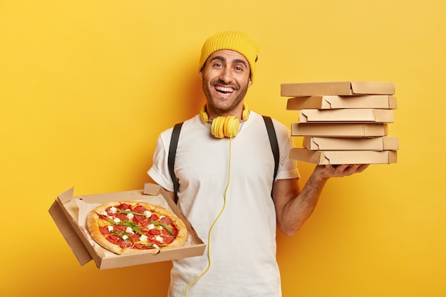 Foto grátis conceito de entrega. o vendedor de pizza homem segura uma pilha de caixas de papelão, mostra um fast food saboroso em um recipiente aberto, trabalha como mensageiro, usa chapéu amarelo e camiseta branca, usa fones de ouvido para ouvir áudio.